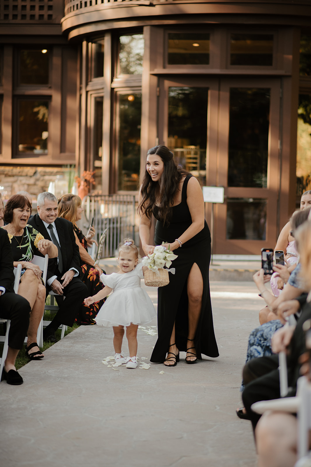 Toddler flower girl walking down the ceremony aisle guided by a bridesmaid in a black dress.